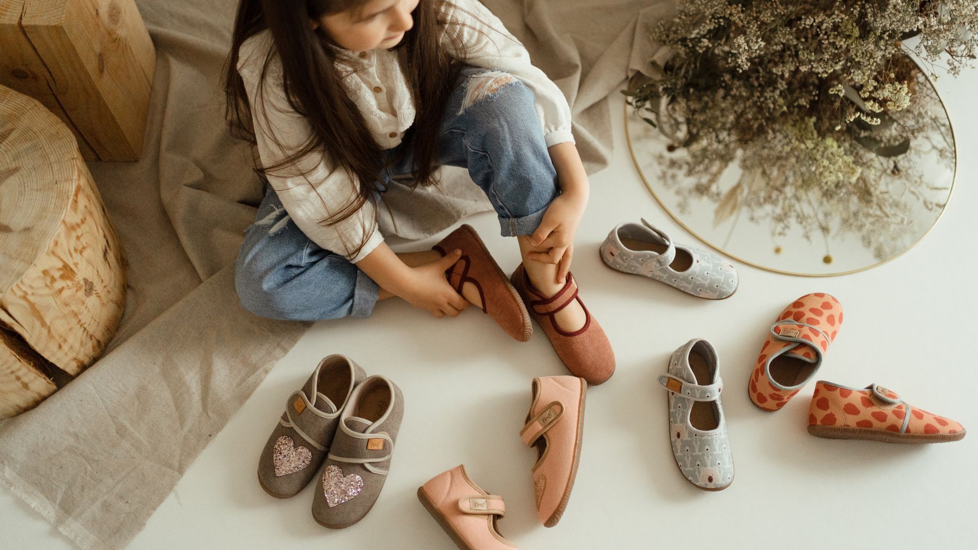 Kapcie dziecięce Emel – dziewczynka siedzi na podłodze wśród różnych modeli ręcznie robionych kapci z naturalnych materiałów, wygodnych i kolorowych.