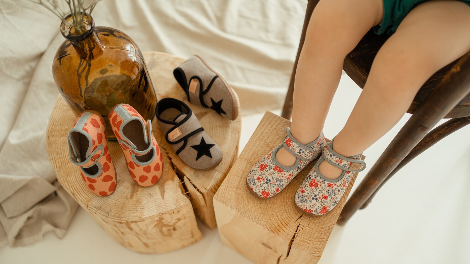 Mała dziewczynka ma na nogach ręcznie wykonane kapcie Emel, obok widoczne dwie inne pary: szare z gwiazdą i pomarańczowe w truskawki, pokazujące różnorodność wzorów.