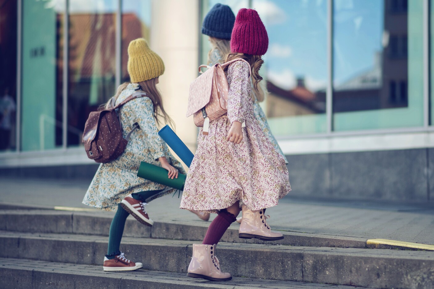 Trzy dziewczynki w skórzanych bucikach Emel wchodzą po schodach, niosąc w rękach brystole, gotowe na kreatywne zajęcia.