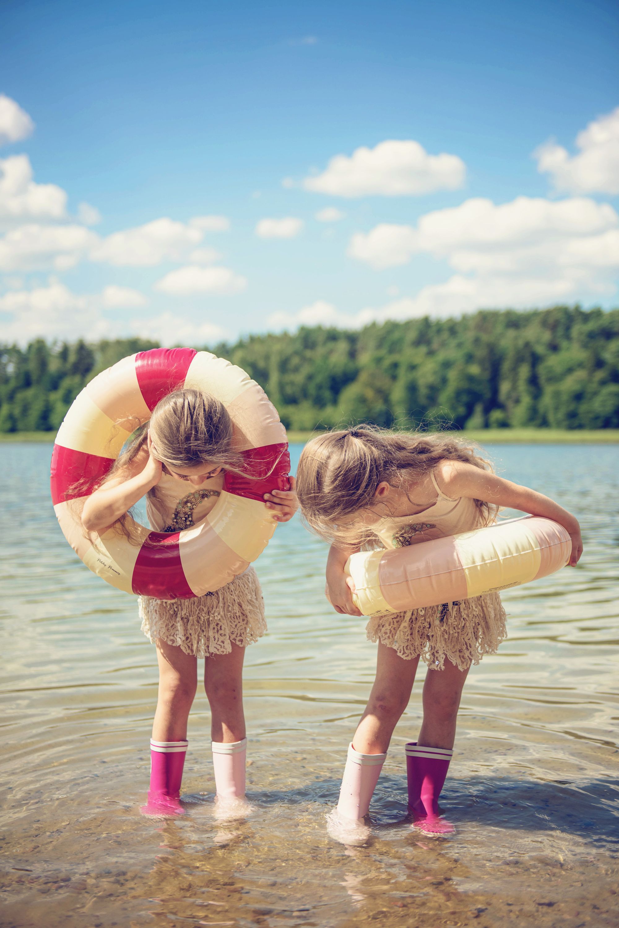 Dziewczynki ubrane w kalosze i koła ratunkowe stoją w jeziorze, z tyłu za nimi jest las i chmury