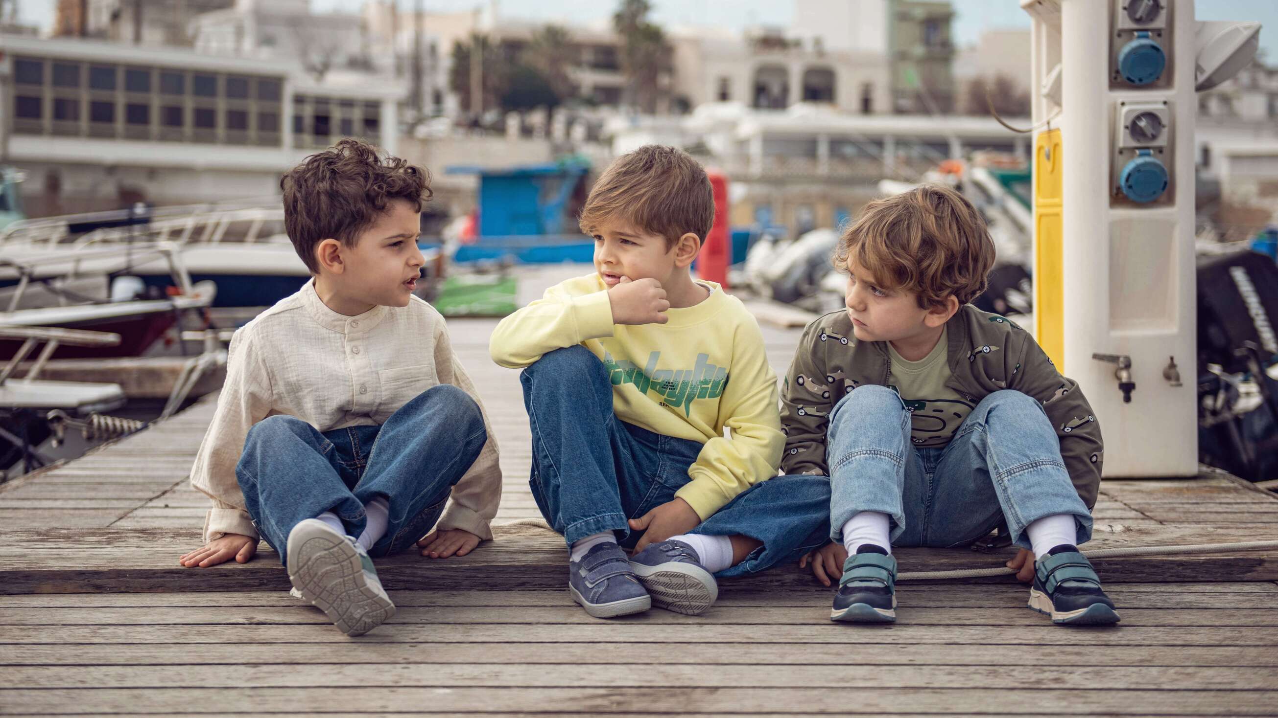 Trzech chłopców siedzi na drewnianym pomoście, mają na sobie skórzane trampki dziecięce firmy Emel.